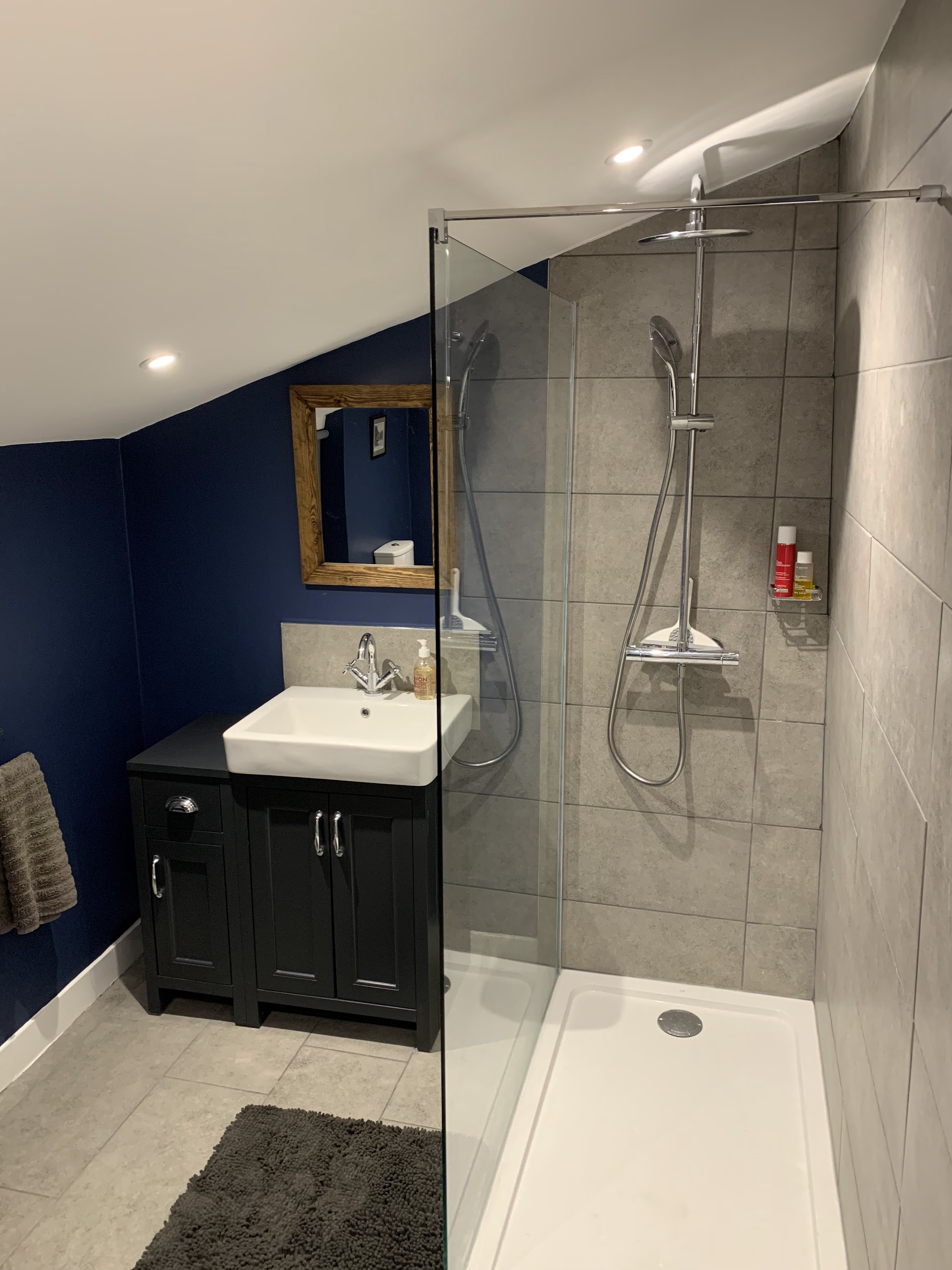 Finished shower room under a sloping ceiling, walk-in glass screen, grey tiles and a dark vanity unit